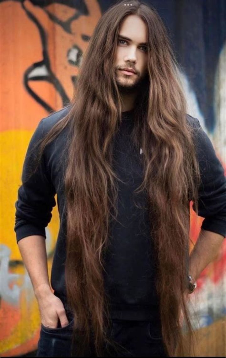 a man with long hair standing in front of a pink background wearing a